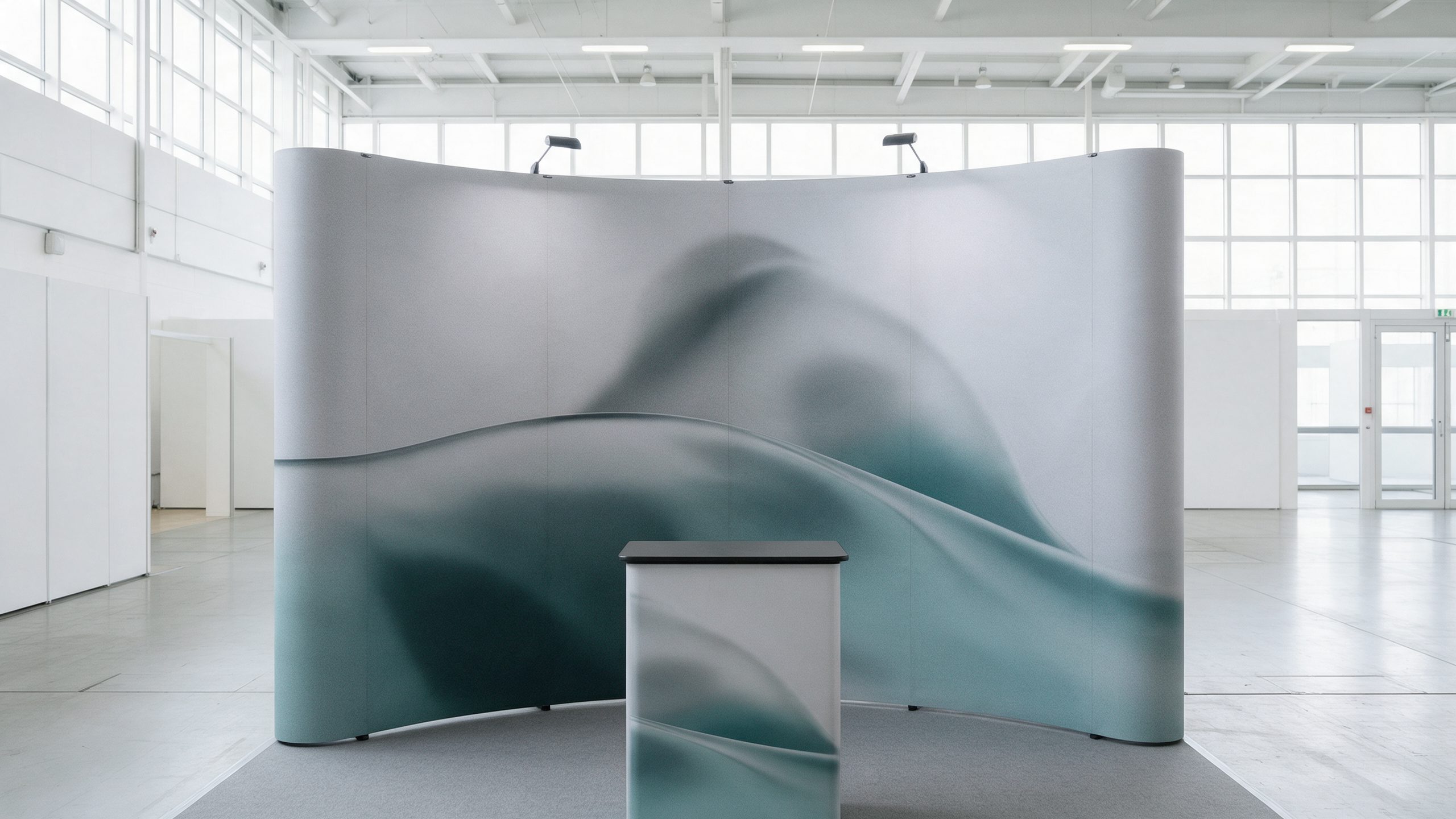 A curved fabric pop-up display wall with plain abstract grey and teal graphics and a small counter in front, in a bright exhibition hall